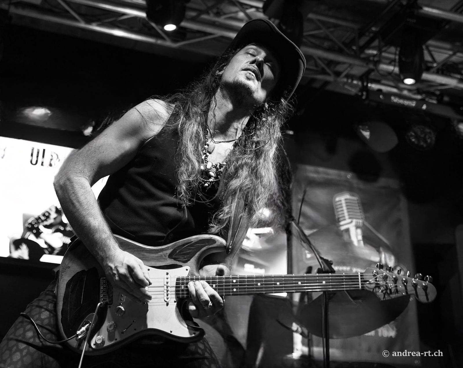 Black and white photo of a passionate rock guitarist with long hair and a hat performing live on stage, captured mid-solo under dramatic lighting – © andrea-rt.ch