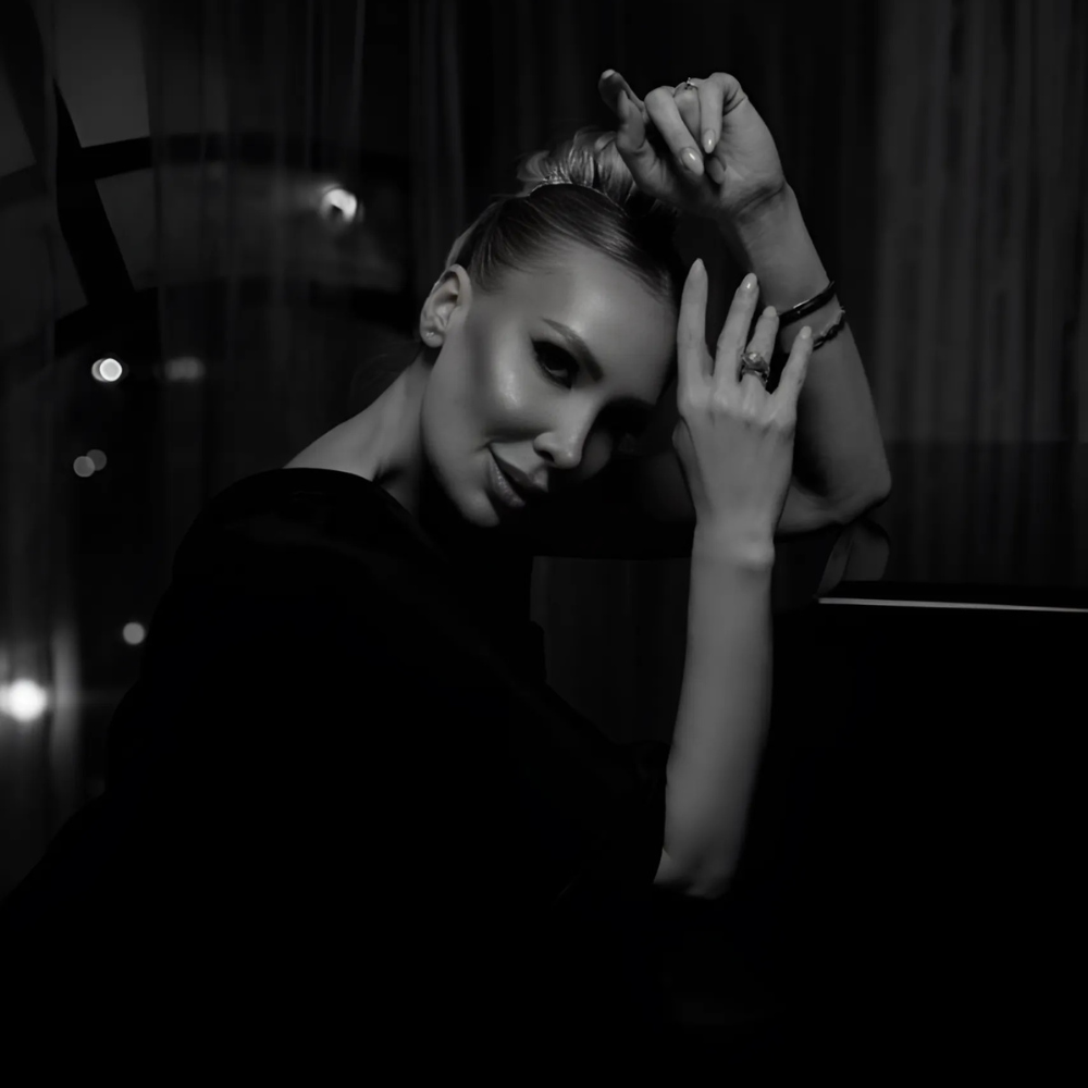 Black and white image of a woman with a sleek updo, gently resting her hands on her head while gazing towards the camera with a confident smile. Soft, blurred lights are visible in the background, creating an elegant and intimate atmosphere.