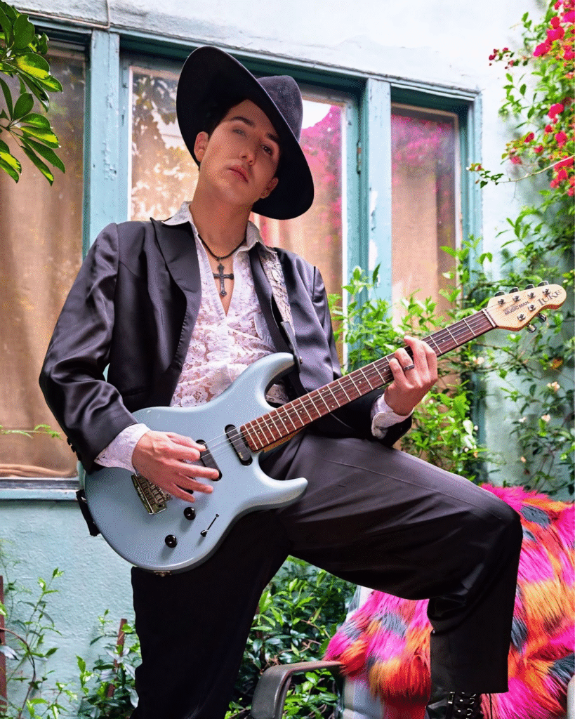 "Musician wearing a black suit and wide-brimmed hat, holding a light blue electric guitar, standing in a vibrant outdoor space with greenery and a colorful background."