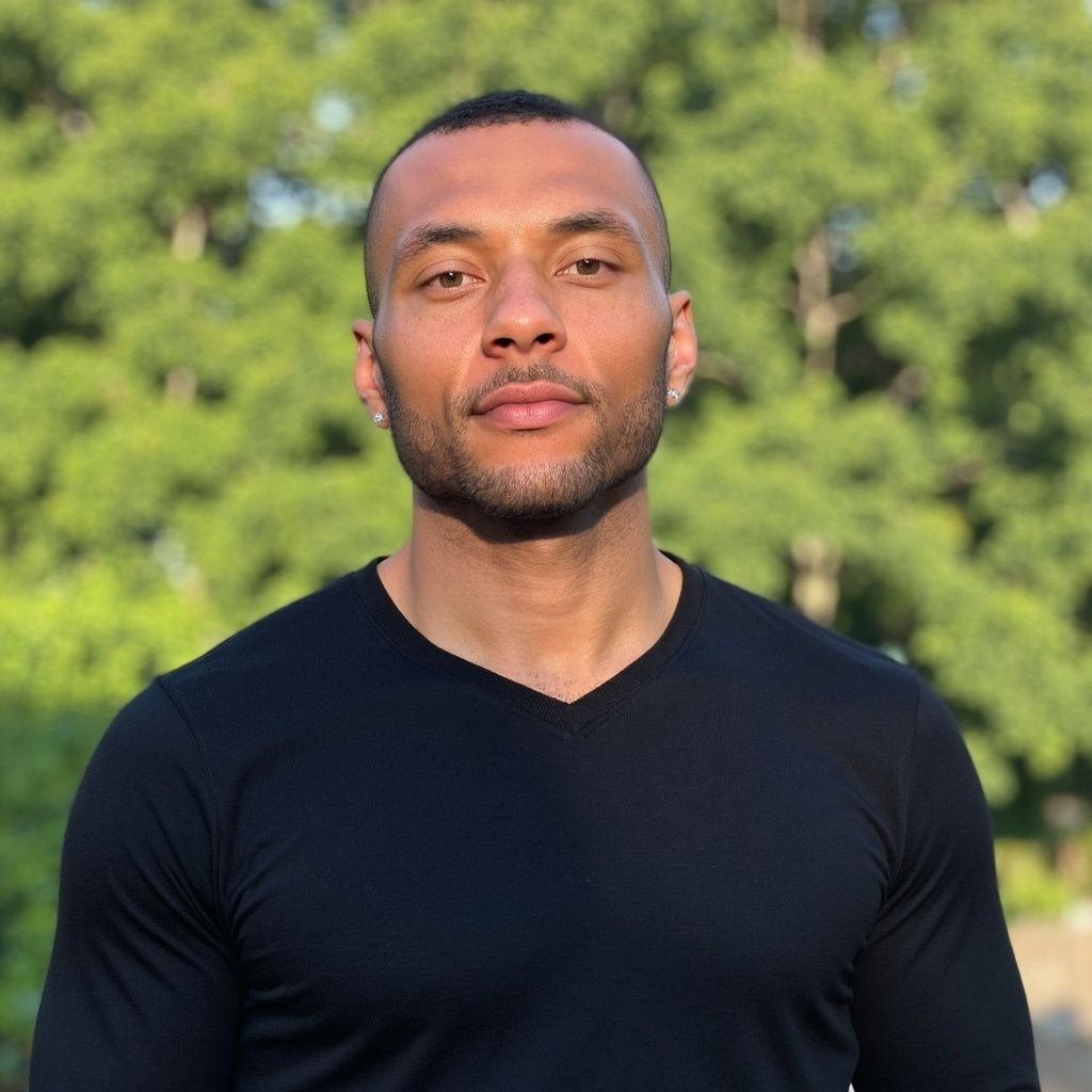 Portrait of a young man with short hair, wearing a black t-shirt, standing outdoors with a blurred green background.