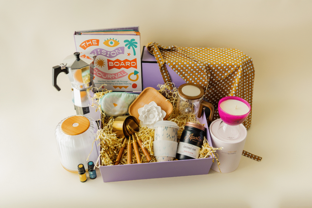 "A curated gift box featuring a Vision Board Journal, moka pot, essential oils, a scented candle, and various artisanal items like wooden spoons, tea cups, and a cozy fabric pouch."