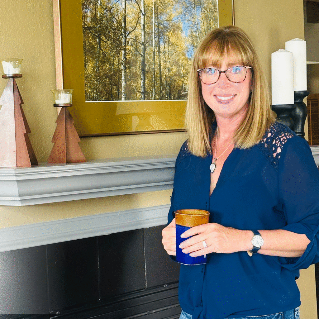 "A woman standing in a cozy living room, smiling and holding a cup. She has light brown hair, glasses, and wears a navy blue shirt with lace detailing. The background features a fireplace and modern decor."