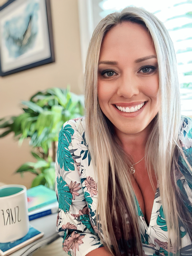 "Close-up of a woman with long blonde hair, wearing a tropical-patterned shirt, sitting at a desk with a cup labeled 'SURRENDER' and plants in the background."