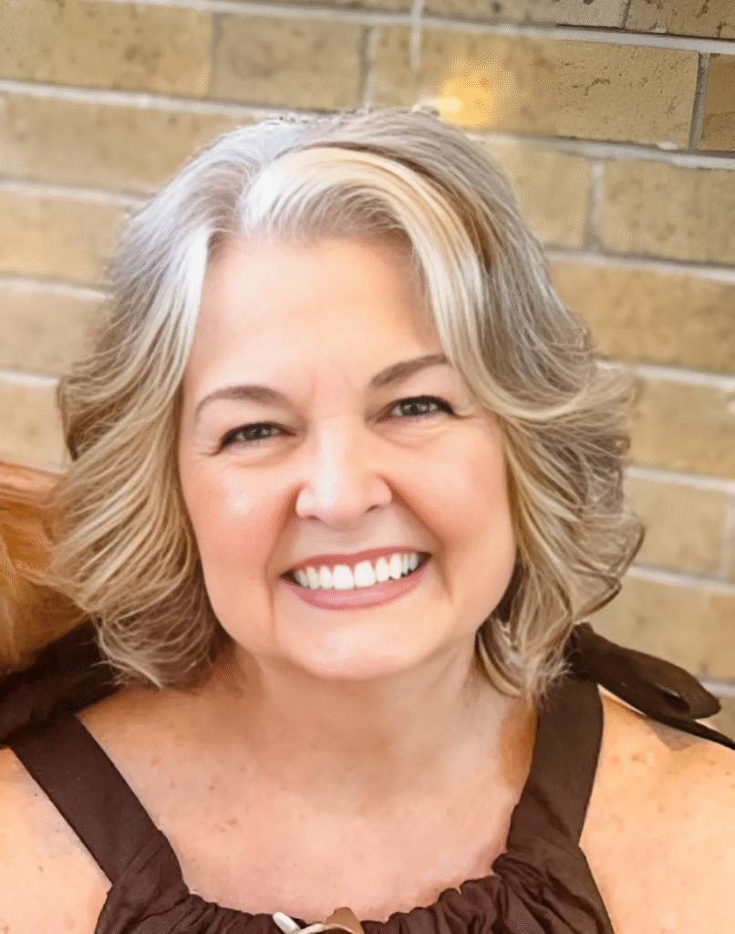 "Close-up portrait of a smiling woman with short, wavy gray hair wearing a brown top. She is smiling warmly at the camera, with a soft background of beige bricks."