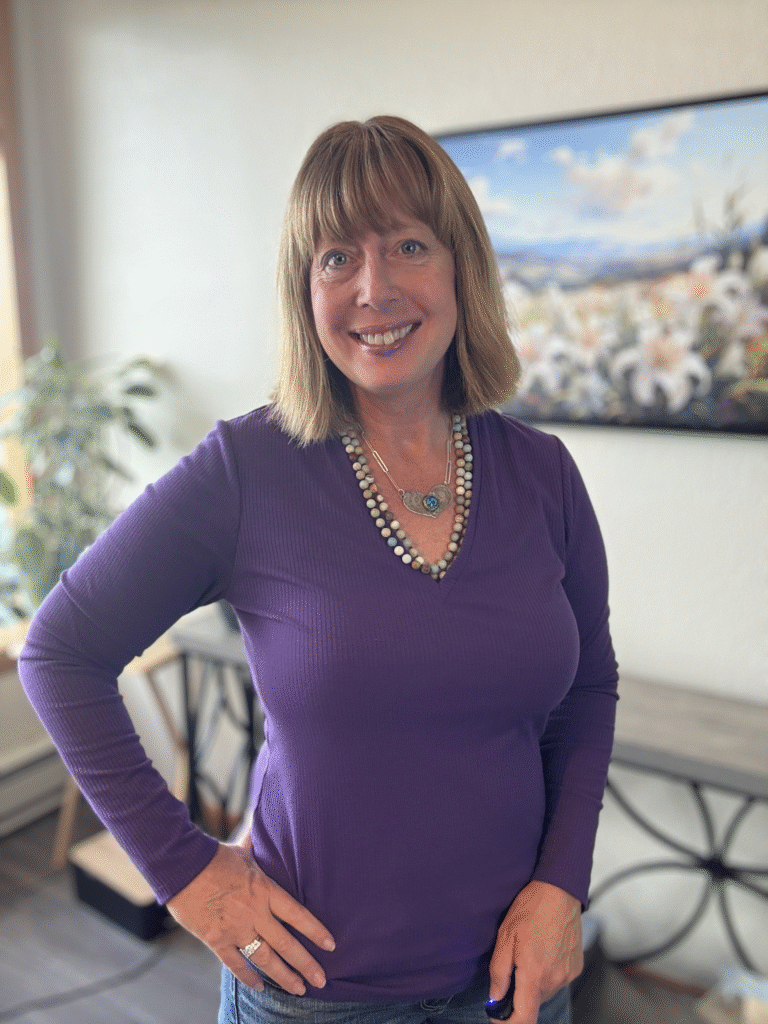"A woman in a purple long-sleeve top, smiling and standing confidently with her hand on her hip. She is wearing a colorful necklace and rings. The background features a bright room with plants and a mountain painting."