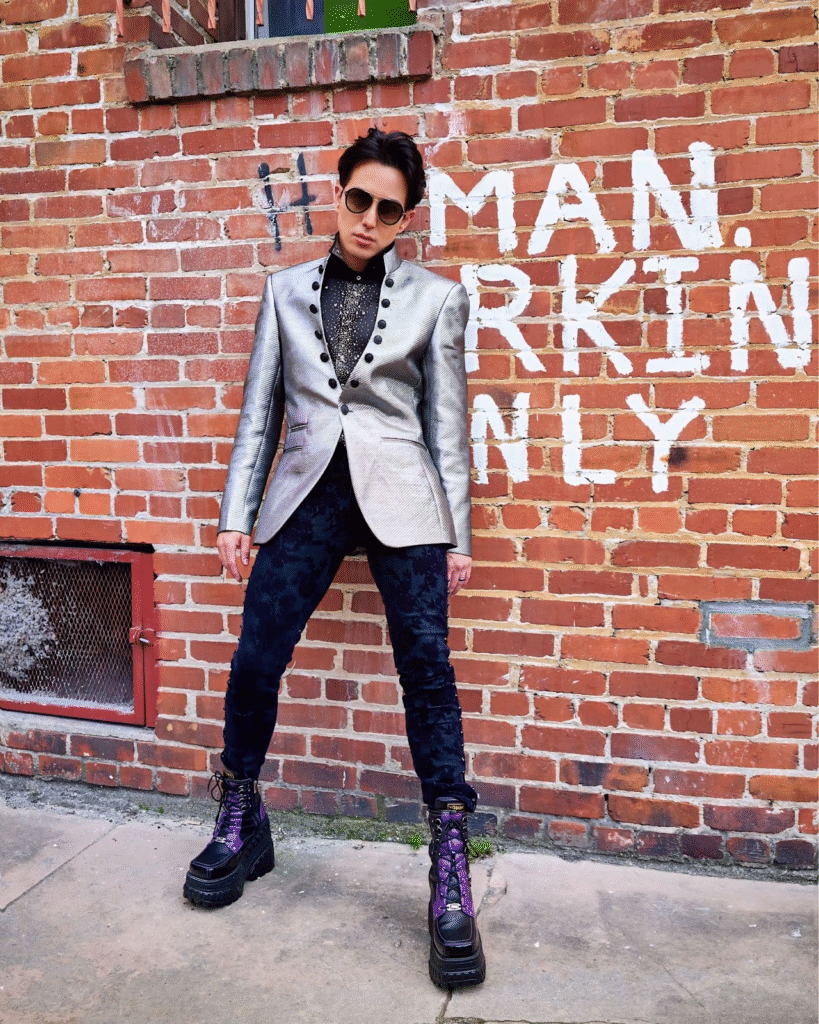 "Fashionable musician wearing a metallic silver blazer, black shirt, and chunky boots, posing in front of a brick wall with the words 'MAN. PARKIN ONLY' spray-painted on it."