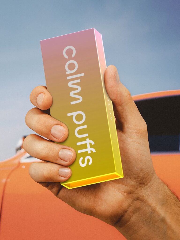 A person holding a brightly colored Calm Puffs box, with a gradient design shifting from pink to yellow, against a background featuring an orange car and blue sky.