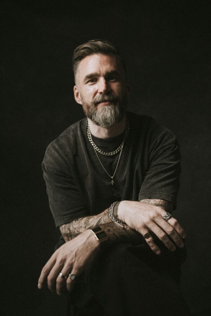 Portrait of Matt Jacob sitting against a dark background, wearing dark clothes and jewelry, smiling slightly.
