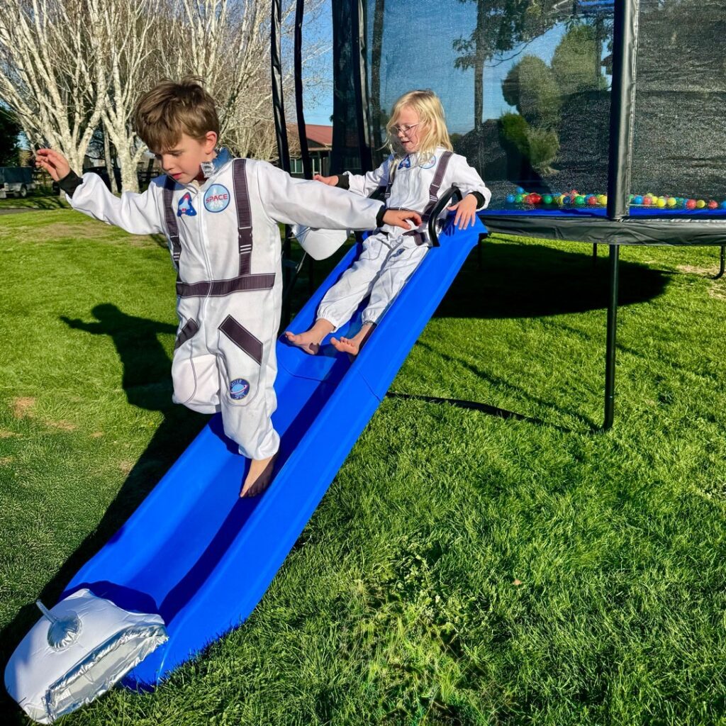 Two kids in astronaut suits playing on blue Rocket Slide attached to SpaceJump trampoline.