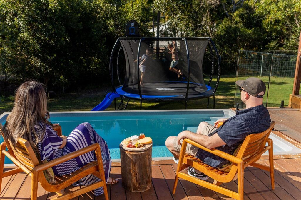 Parents relaxing by pool watching kids jump on SpaceJump trampoline.