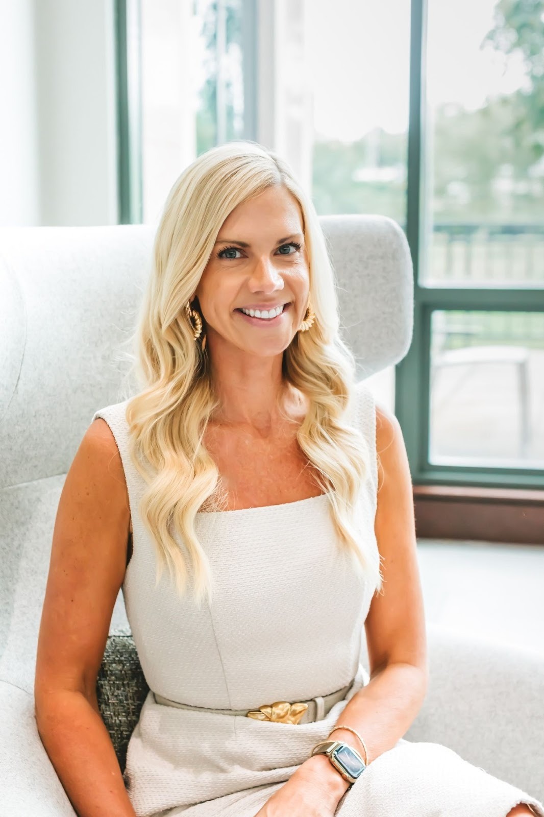 Smiling woman sitting comfortably in a chair, wearing a light-colored dress and gold accessories.
