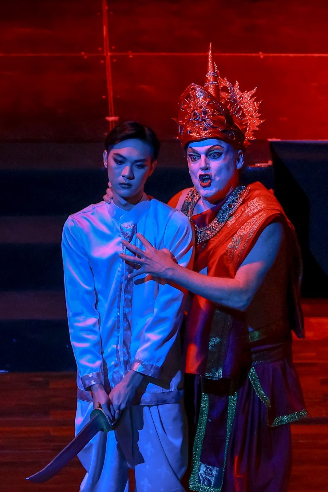 A male and female performer sing during a cultural event, with a choir and orchestra performing in the background.