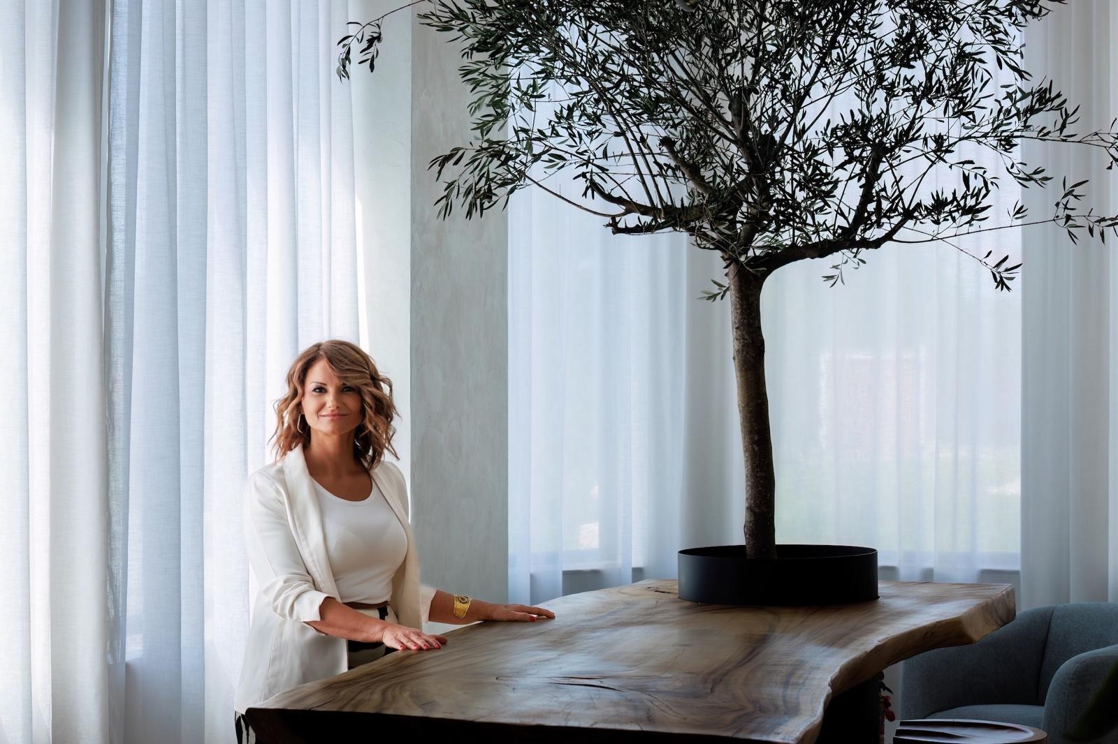 A woman in a white blazer standing next to an olive tree in a modern space.
