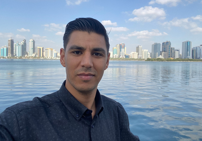 A man standing by the water with a city skyline in the background.