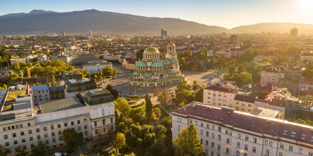 Aerial view of the Alexander Nevsky Cathedral surrounded by city buildings in Sofia, Bulgaria, with mountains in the background at sunset.