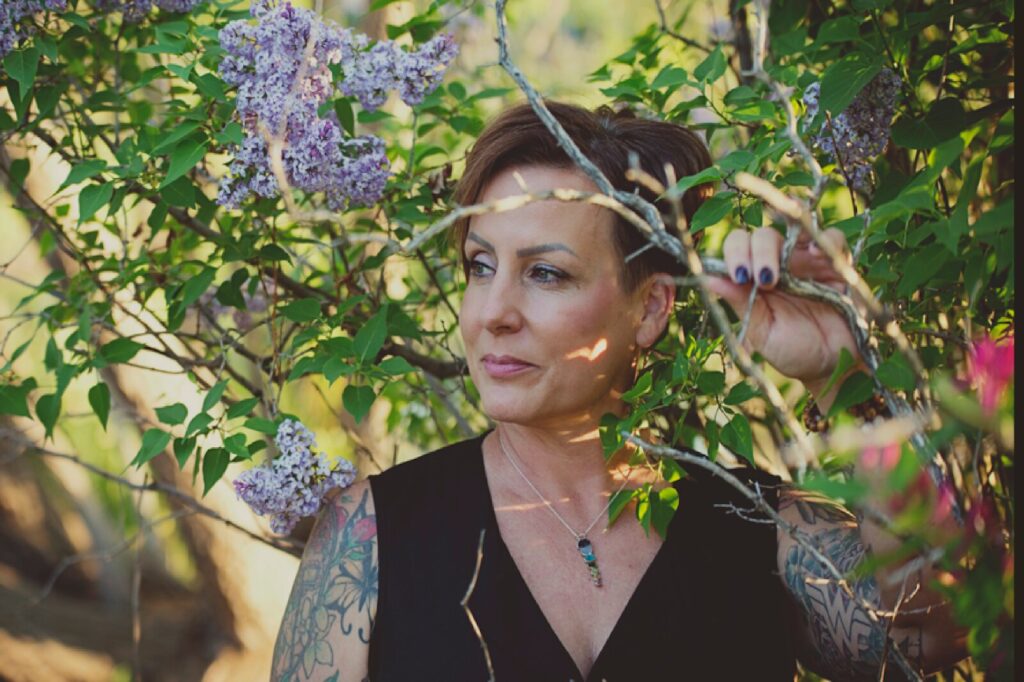 A woman with tattoos in a black top stands among colorful flowers, embodying strength and beauty in a serene setting.