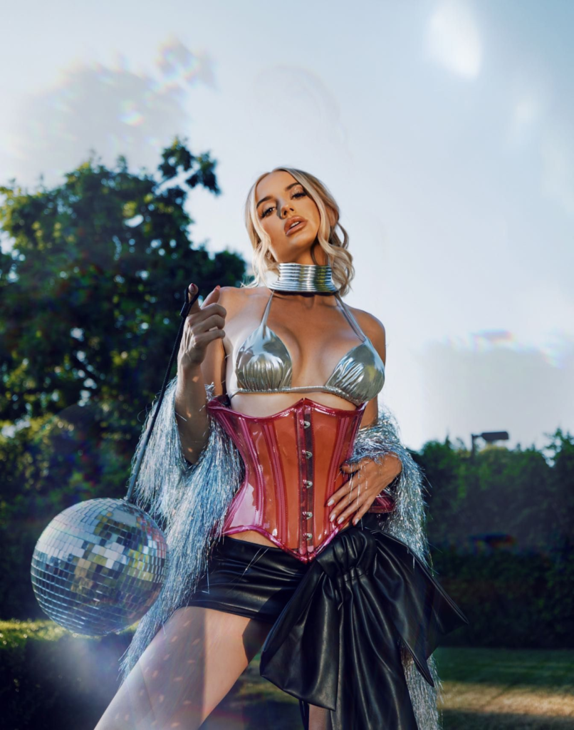 Woman in metallic bikini top and corset holding disco ball, posing outdoors with trees in background.