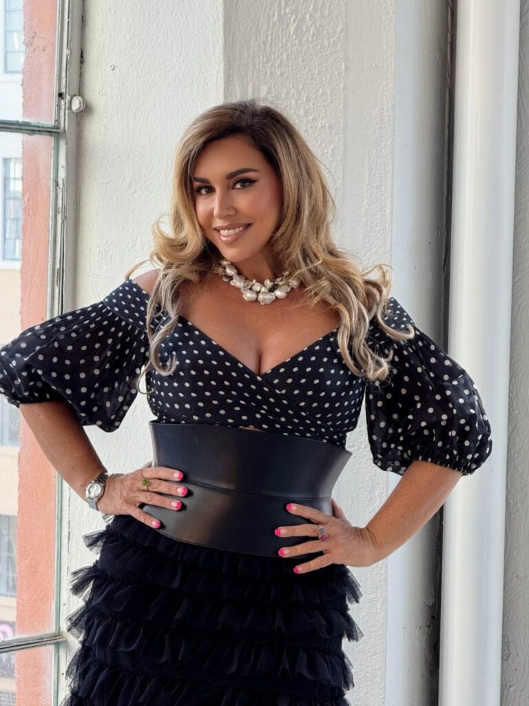 Woman wearing black polka-dot dress and pearl necklace posing by window with hands on hips.