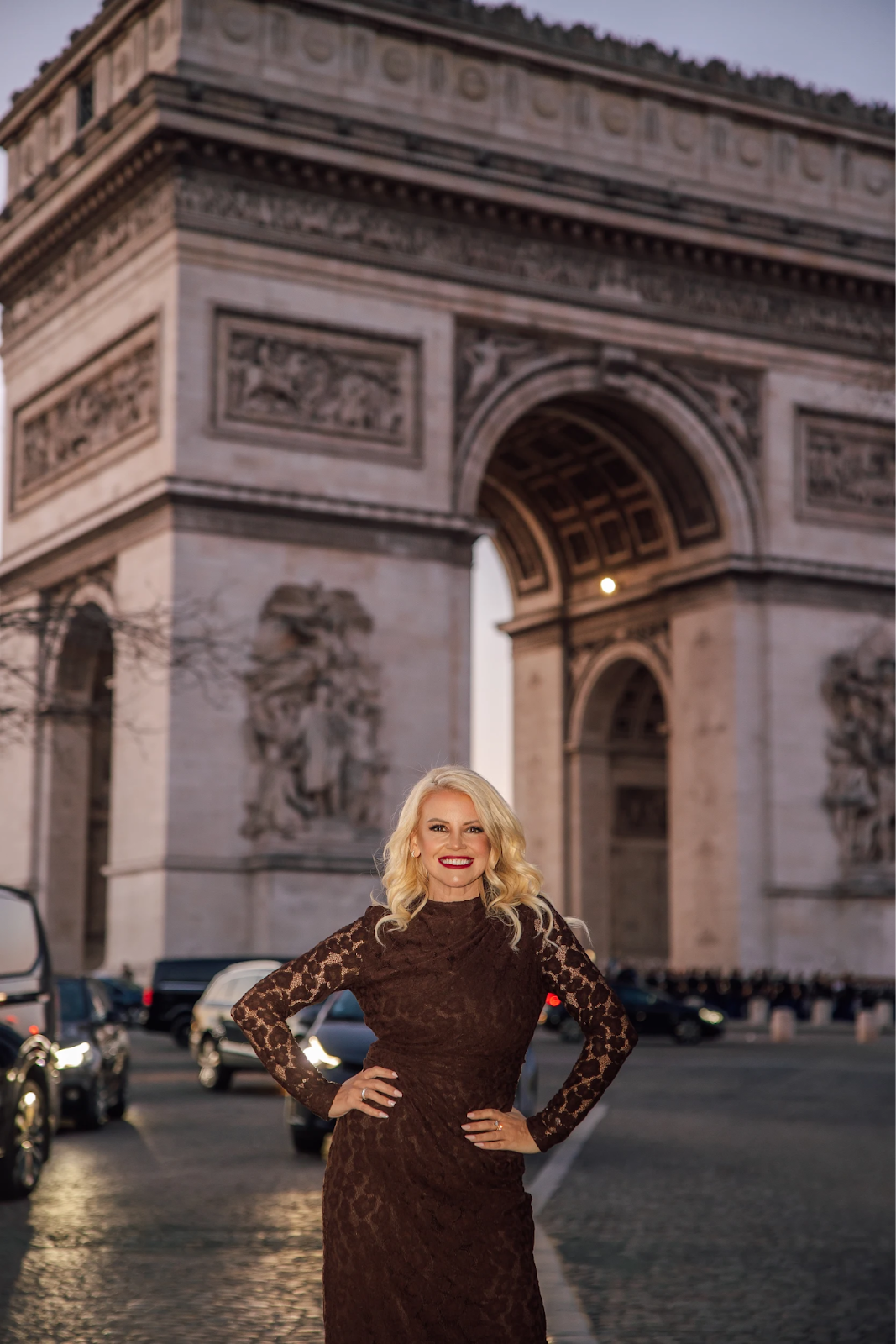 Blonde woman in brown lace dress posing before Arc de Triomphe with evening traffic.