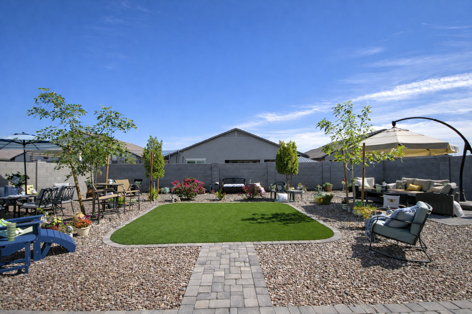 Landscaped Arizona backyard with green lawn, patio seating, umbrellas, trees, and gravel garden under bright blue sky.