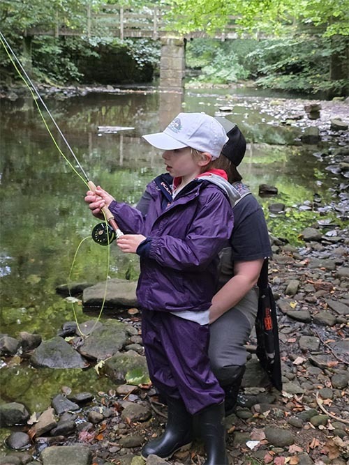 A man and a child enjoy fishing together by a river, surrounded by lush greenery and calm water, sharing a bonding moment.