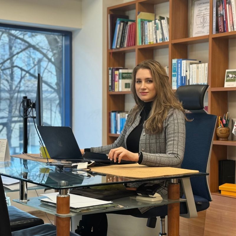 A woman sits at a glass desk with a laptop and papers in a modern office, with bookshelves and a large window in the background.