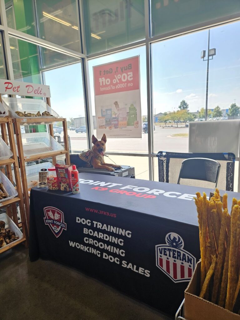 Dog resting on crate at indoor Joint Forces K9 Group booth with training services display.