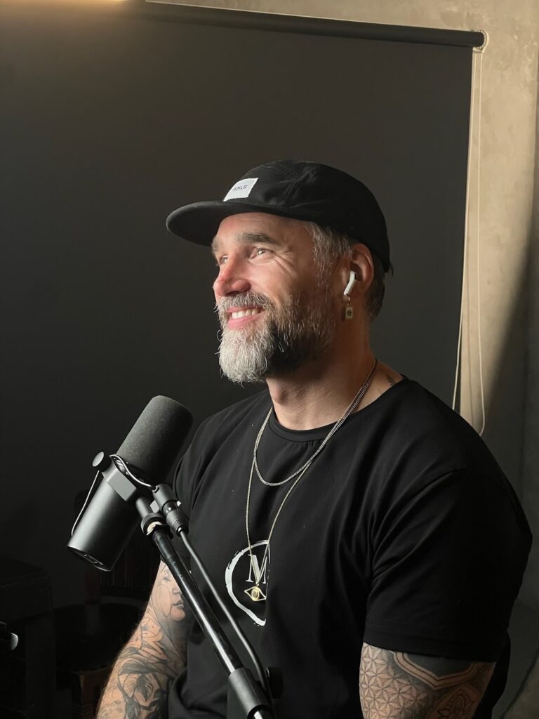 A tattooed man in a hat sits before a microphone, ready to host The MOOD Podcast, exploring deep creative conversations.