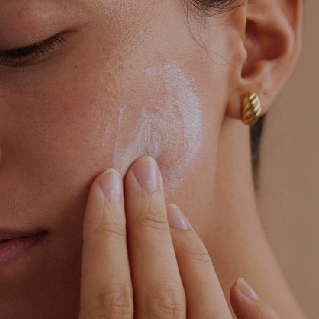 Person applying white skincare cream to cheek with fingers, close-up view.