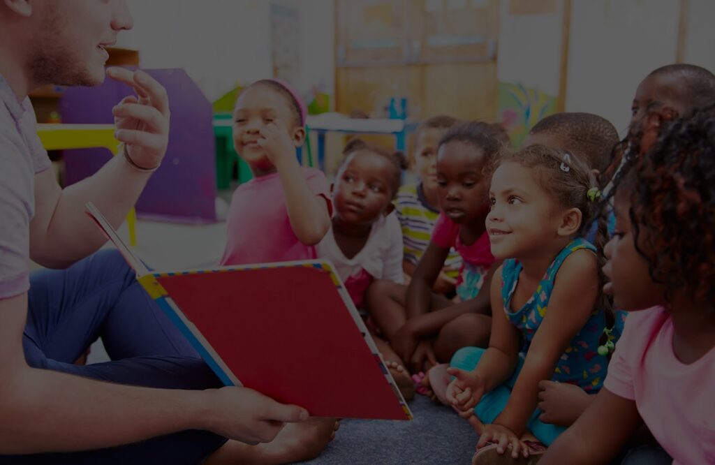 Teacher reading a book to attentive preschool children gathered in a colorful classroom.