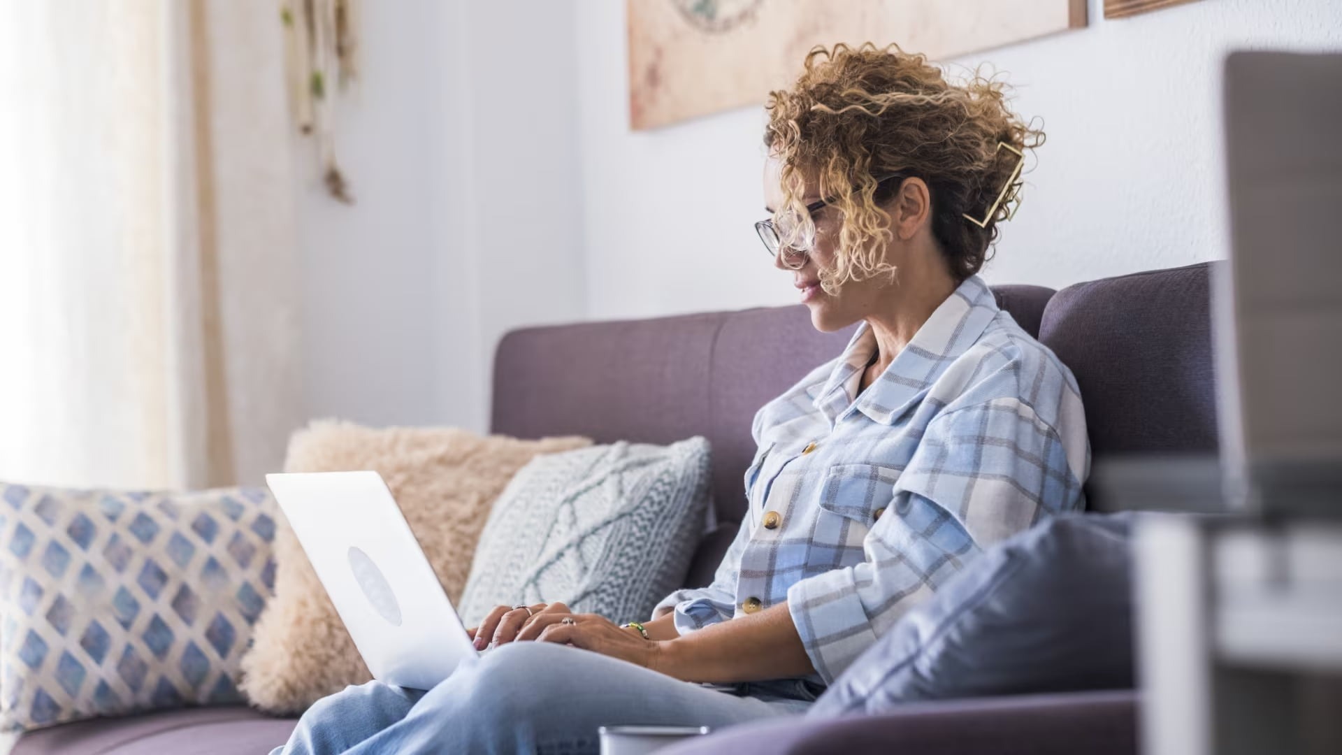 Woman with curly hair and glasses sits on a sofa, focused on her laptop. She wears a plaid shirt, surrounded by cozy pillows and soft light.