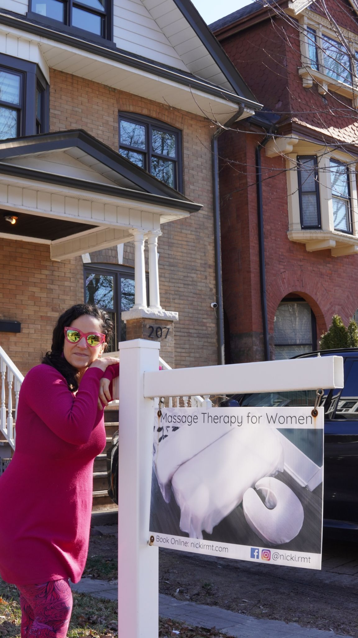 A woman in a pink outfit leans on a white post with a massage therapy sign outside a brick house. She wears reflective sunglasses and smiles.