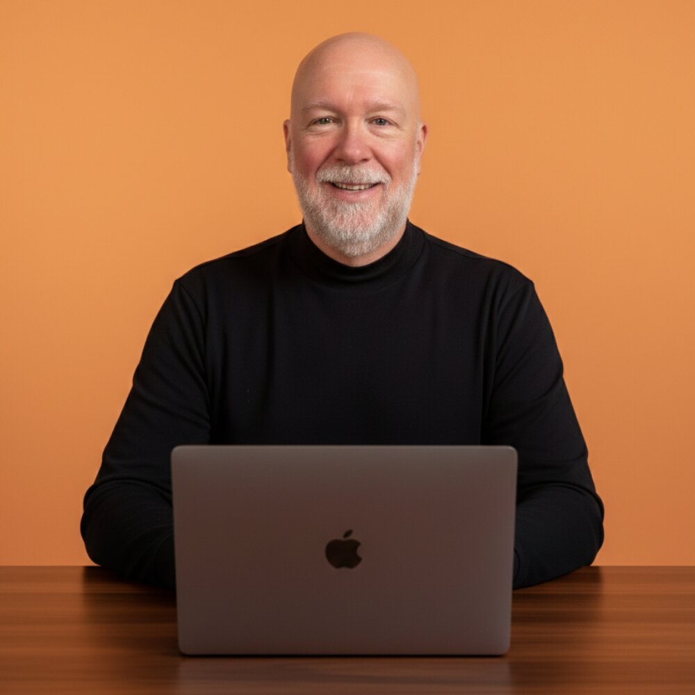 A bald man sits at a table with an Apple laptop, reflecting on his journey from military leader to financial planner for veterans.