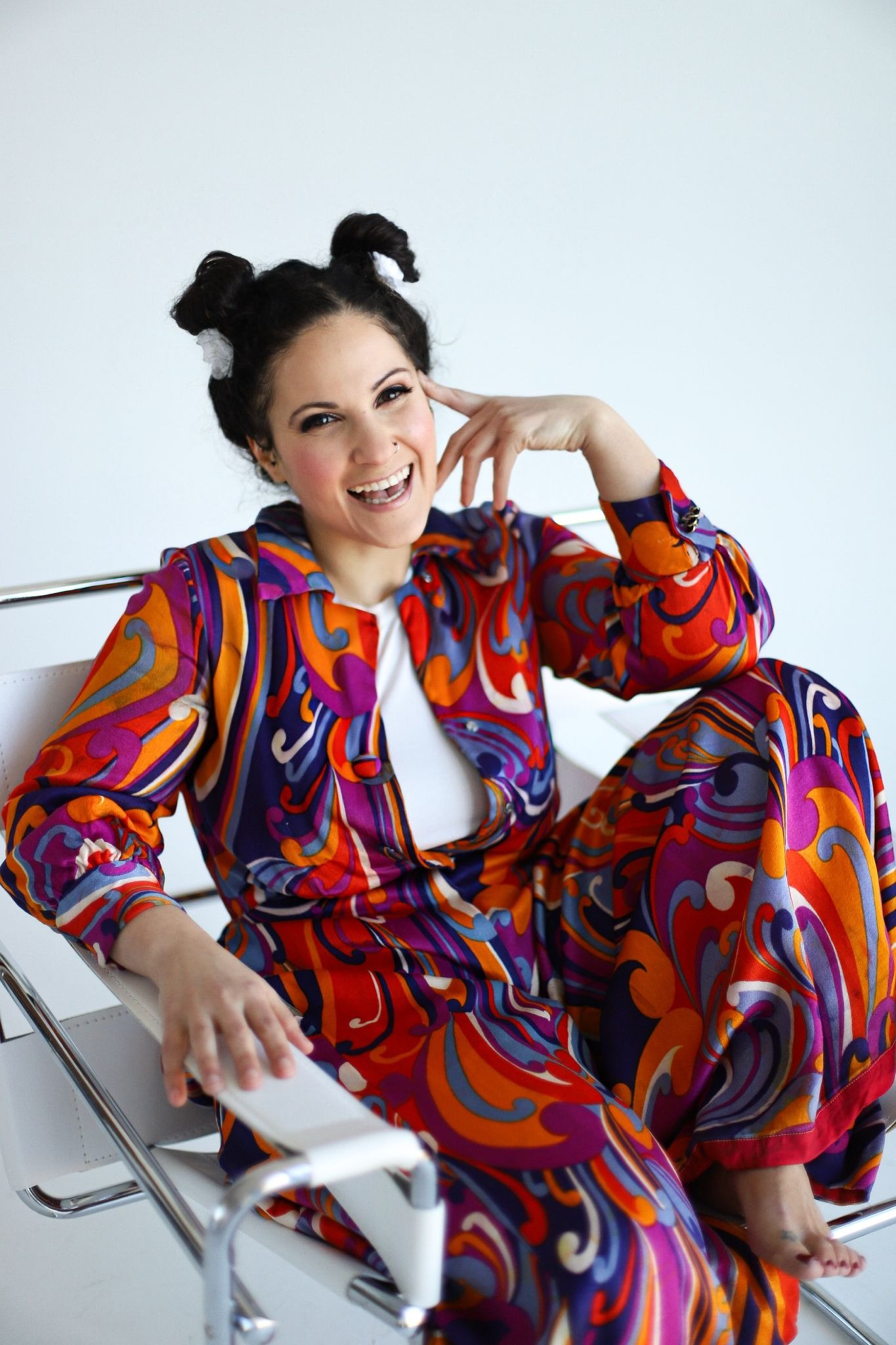 Smiling woman with dark hair in playful buns, wearing a colorful, vibrant shirt with swirling patterns, poses with hand touching her face. Bright and cheerful mood.