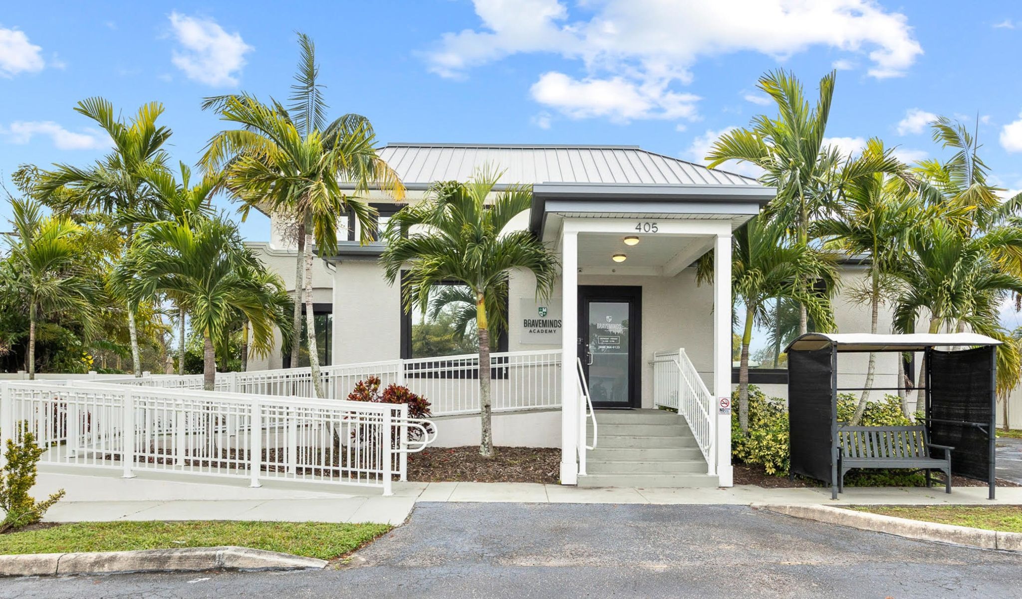A bright, sunny day frames a small white building with a metal roof, surrounded by tropical palm trees. A wheelchair ramp and bench enhance accessibility.