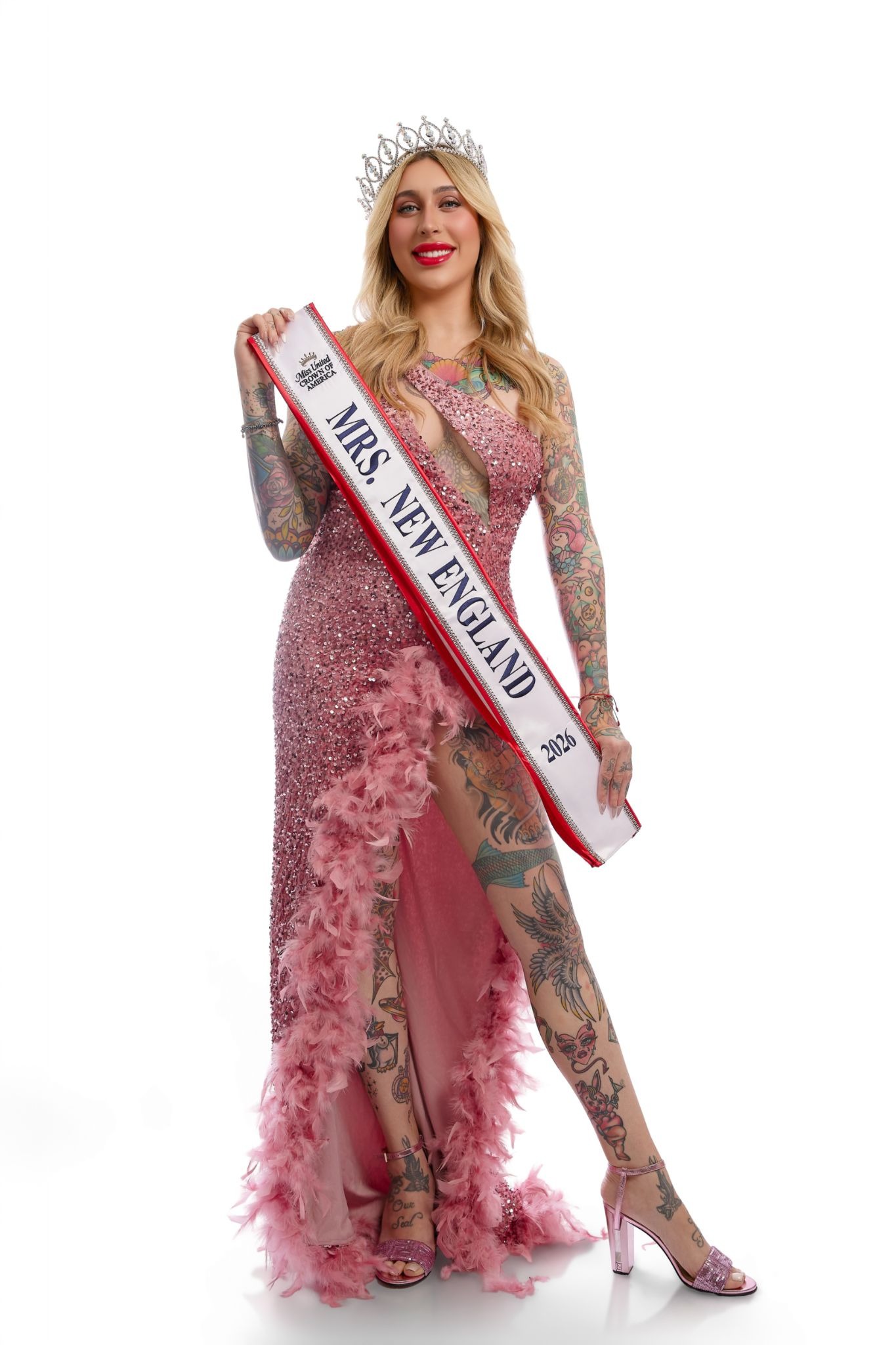 A woman in a sparkling pink gown, adorned with tattoos, wearing a crown and holding a “Mrs. New England 2023” sash, smiles confidently.