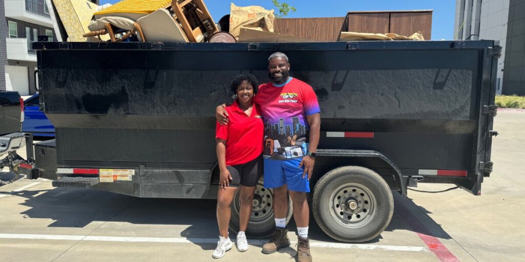 A smiling man and woman stand arm-in-arm in front of a large black trailer filled with furniture. They appear happy and relaxed on a sunny day.