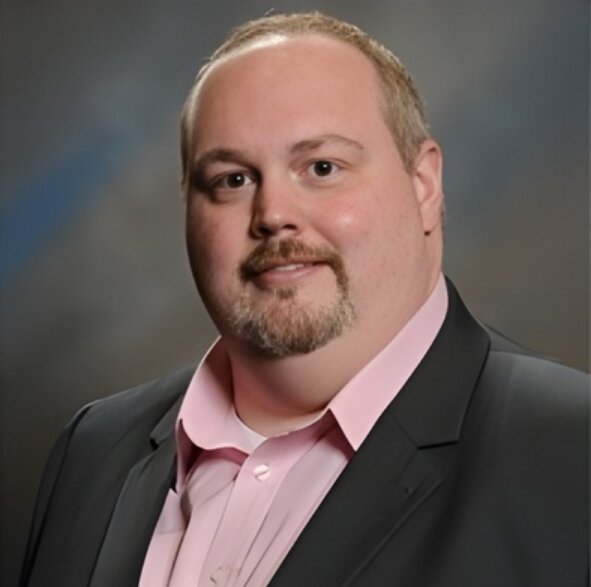 A person with short, light hair and a goatee, wearing a pink shirt and dark suit, smiles subtly against a blurred gray background. The tone is formal yet approachable.