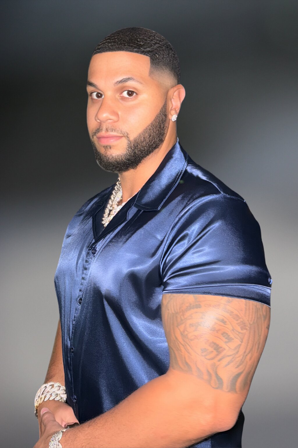 Man in a glossy navy shirt poses confidently. He has a trimmed beard, diamond earrings, an intricate arm tattoo, and a chain bracelet, exuding elegance and strength.