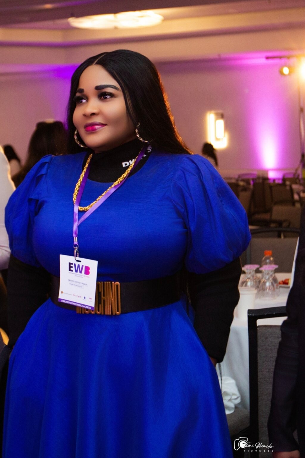 A woman in a vibrant blue dress with puffy sleeves stands confidently at an event. She wears a Moschino belt and a name tag, surrounded by a softly lit room.