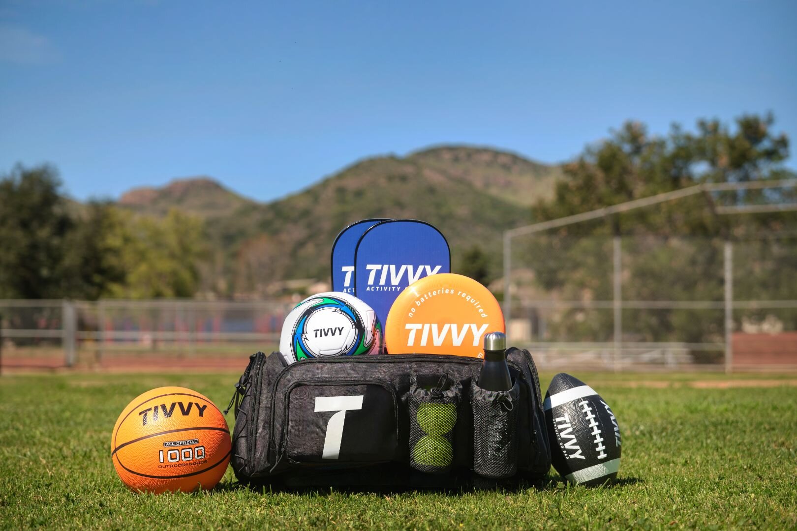 A sports bag with various equipment, including a basketball, soccer ball, football, paddles, and cones, sits on grass against a backdrop of mountains and a clear blue sky.