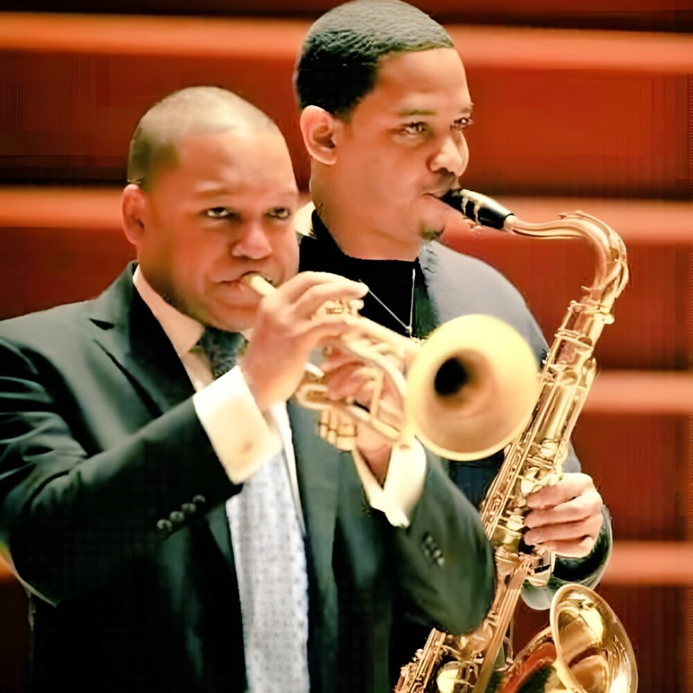 Two musicians in suits play brass instruments passionately on stage, one with a trumpet and the other with a saxophone, creating a focused, harmonious atmosphere.