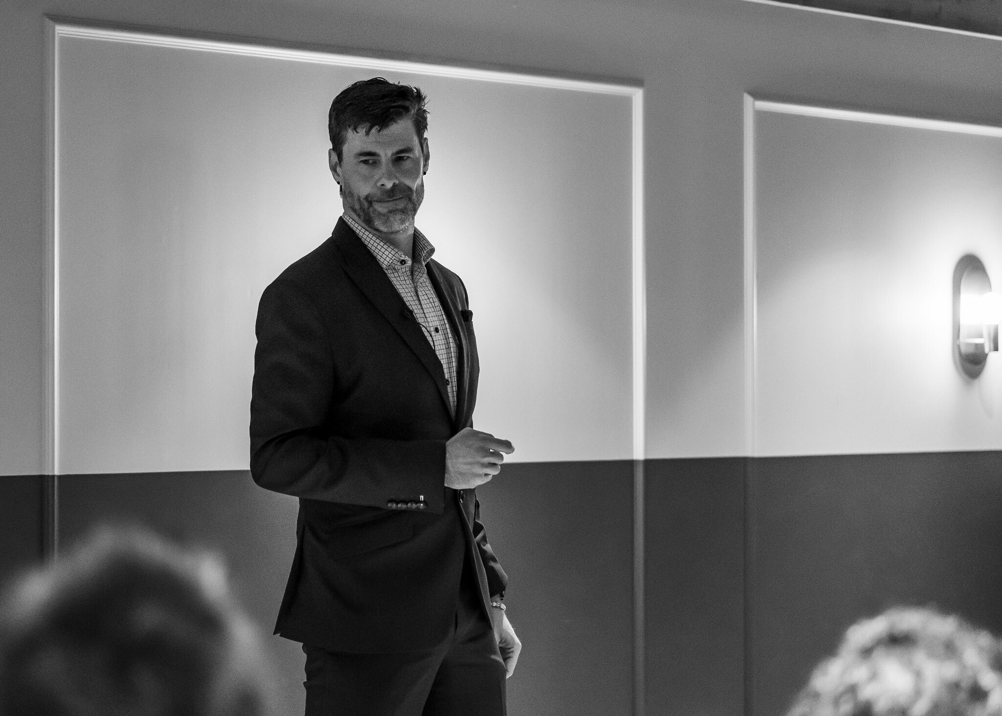 A man in a suit stands confidently, speaking to an unseen audience in a dimly lit room. The atmosphere is professional and focused.