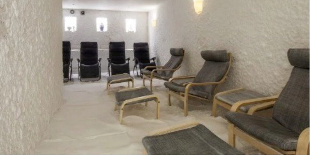 A serene salt room with white, textured walls and floor. Several cushioned lounge chairs and footrests are arranged, creating a peaceful, relaxing atmosphere.