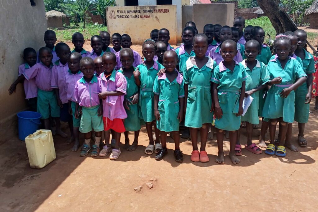 A group of children in uniform stand together outdoors, smiling. They wear green and purple outfits. A school sign is visible in the background.
