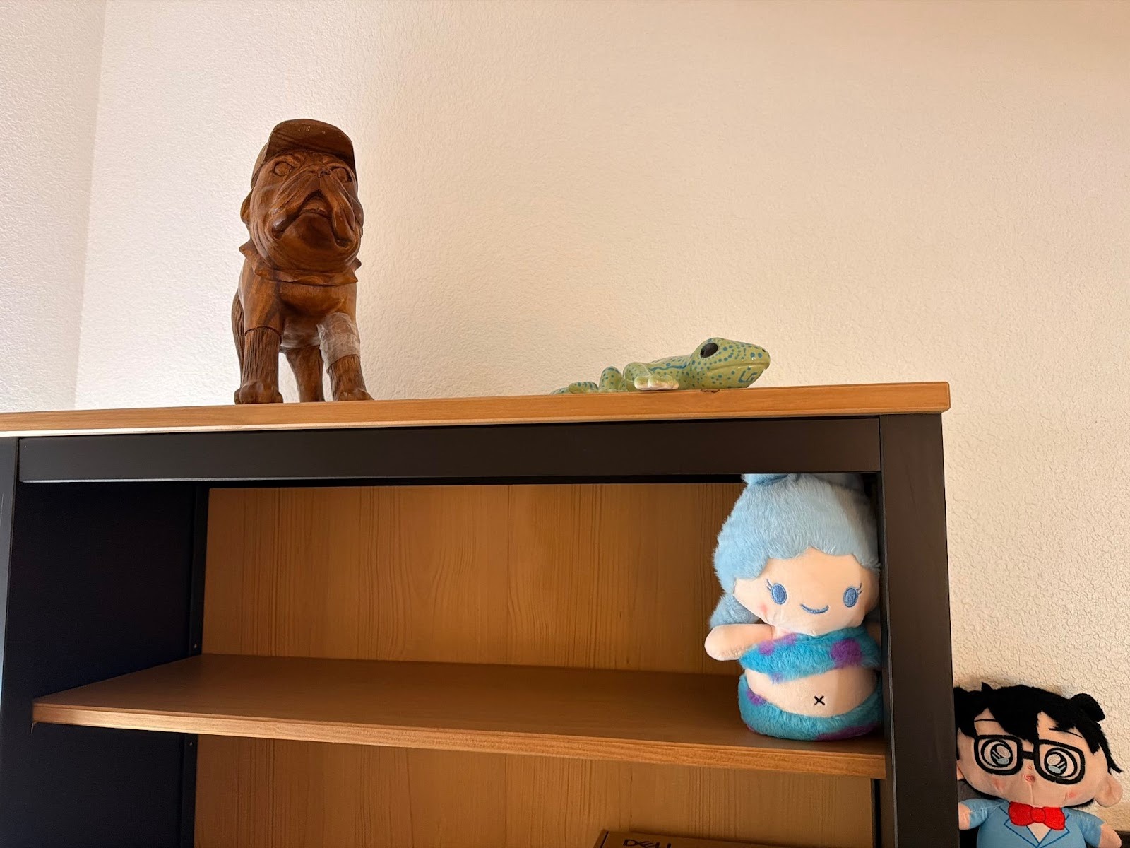 A wooden shelf with a carved wooden dog statue and a ceramic frog on top. Below are plush toys, including a blue-haired character and a figure with glasses.