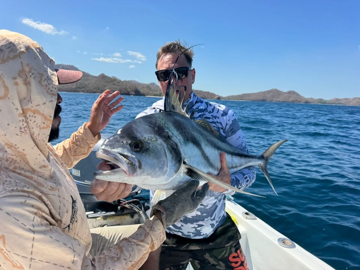 Two fishermen holding large roosterfish on boat with coastal hills in background.