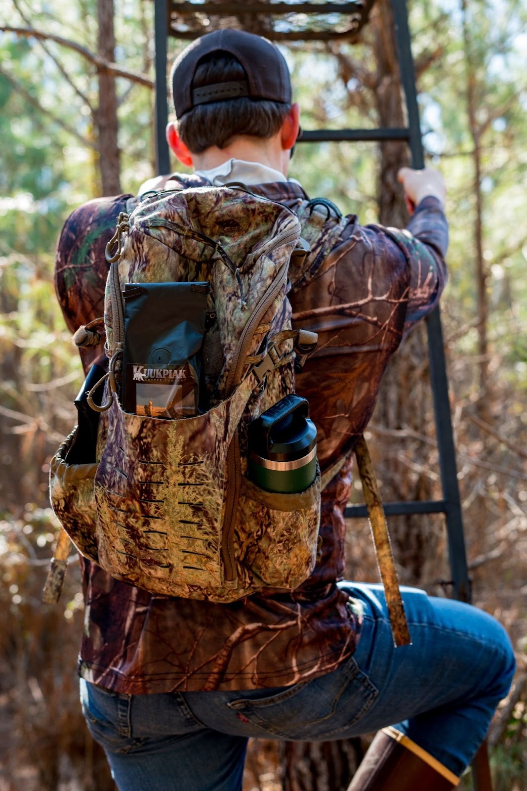 Man in camouflage gear climbs a tree stand ladder in a forest. He carries a camo backpack with a thermos and gear. Sunlight filters through trees.
