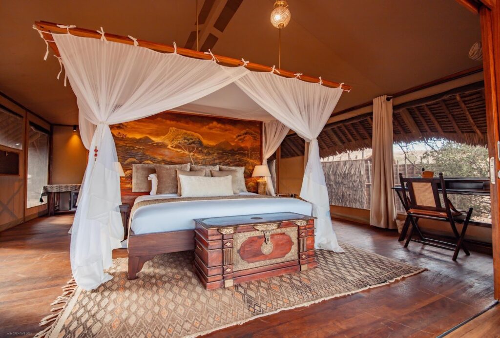 A luxurious safari lodge bedroom with a four-poster bed draped in white netting, warm earthy decor, and a view of nature through open curtains, creating a serene ambiance.