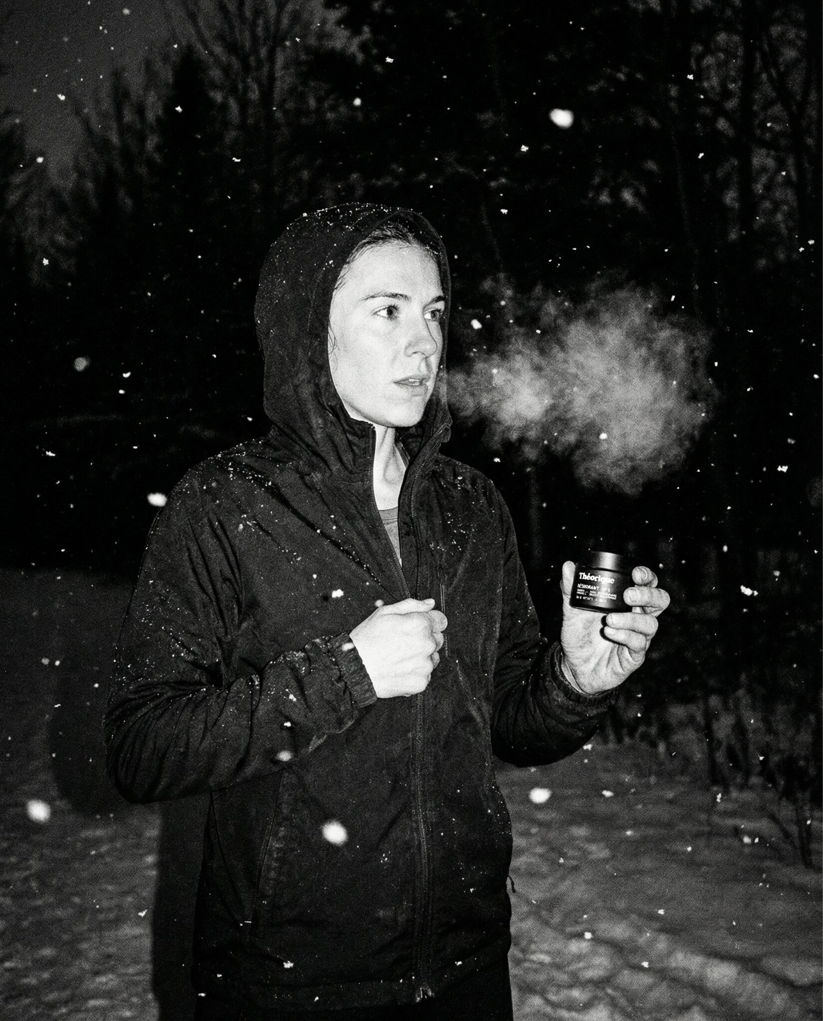 Black-and-white image of a person in a hooded jacket standing in falling snow at night, holding a small jar, visible breath suggesting cold air.